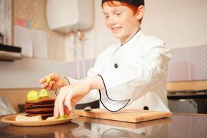 Kind Junge im Koch Kostüm bereitet vor Schokolade Pfannkuchen im Küche im Cafe. schön rutschen von Pfannkuchen mit Obst und Schokolade Nahansicht foto