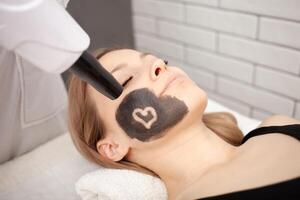 Kosmetikerin tut Verfahren von Kohlenstoff Peeling von Gesicht. schwarz Maske, Laser- Haut Reinigung. modern Behandlungen. schneidet Herz foto
