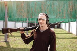 ein Mann im schützend Brille und Kopfhörer. ein Pumpwirkung Feuerwaffe Schrotflinte. Reifen draußen. Grün Gras, Schutz zum Sport Schießen. Hobby. foto