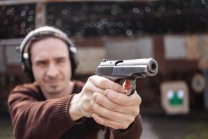 ein Mann schießt ein Pistole, Zielen beim das Ziel. ein Mann tragen schützend Kopfhörer. ein Mauer und ein Dach mit Kugel Löcher. draussen Schießen Angebot foto