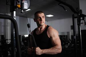 ein Sport Athlet Trainer ein muskulös Mann tut ein Übung auf ein Simulator im Fitnessstudio. Porträt, niedrig Taste, Dunkelheit. baut oben Arm Muskeln foto