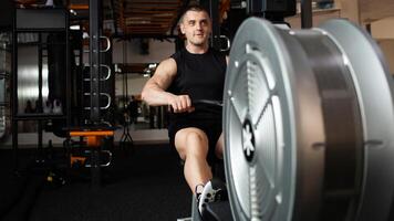 gut aussehend männlich Trainer sitzt auf ein Simulator im Fitnessstudio. Nachahmung Rudern, Boot mit Ruder, zum Ruderer. lächelnd, Glücklich, Sport aktiv gesund Lebensstil, Innen- Konzept, Coaching und Individuell Unterricht. Banner foto