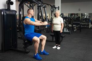 gut aussehend jung Trainer Mann zeigt an Senior Frau Wie zu richtig ausführen Übungen auf das Simulator im das Fitnessstudio. glücklich Menschen lächeln, genießen, glücklich. aktiv Sport gesund Lebensstil drinnen. foto