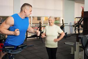 ein schön jung Trainer zeigt an das alt Frau Wie zu tun das Übung auf Simulator. Menschen lächeln, sind Glücklich, Show Klasse, Daumen. super. Sport aktiv gesund Lebensstil Konzept. Senior foto