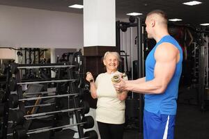 ein alt Frau und ein jung männlich Trainer tun Übungen im Fitnessstudio. Sie Shake ihr Muskeln mit Hanteln, lächeln, sind glücklich. aktiv, gesund, sportlich Lebensstil, Senior Konzept drinnen. Porträt foto