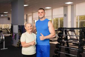 Porträt von ein jung Trainer im Uniform und ein alt Frau im Fitnessstudio. physisch Kultur zum älter Personen. Ausbildung und Individuell Kurse. glücklich Menschen lächeln, Sport ist Cool, super, Daumen hoch. Konzept Senior foto