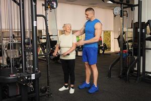 jung gut aussehend Trainer Mann hilft ein Rentner zu tun ein Übung auf das Simulator im das Fitnessstudio. glücklich Menschen lächeln. aktiv gesund Sport Lebensstil, Senior Konzept. foto