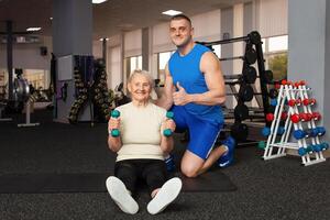 jung männlich Trainer hilft ein alt Frau Übung im das Fitnessstudio. aktiv, sportlich, gesund Lebensstil. Senior Konzept. drinnen. glücklich lächelnd Menschen, Pensionierung Alter. suchen in das Kamera foto