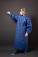 das Arzt zeigt an auf etwas mit seine Hand auf ein grau Hintergrund, ein leeren Raum zum Text. ein Mann im ein Blau Kleid, Hut, Handschuhe, Bahilla, im ein medizinisch Maske. foto