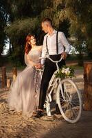 ein schön Paar im das Wald auf ein Weiß Fahrrad mit ein Strauß von Blumen. Hochzeit gehen, ein Frau im ein grau Kleid, ein Mann im ein passen und Hosenträger suchen beim jeder andere foto