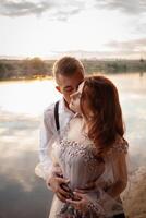 ein schön Hochzeit Paar auf das Flussufer beim Sonnenuntergang. ein Frau im ein grau Kleid mit Blumen, ein Mann im ein passen und Hosenträger Umarmung jeder andere, Liebe. Liebhaber auf ein gehen foto