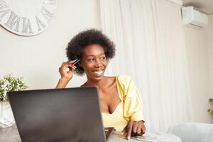 schön afrikanisch amerikanisch schwarz Frau Verwendet ein Bank Karte zu Einkaufen online. Lieferung von Waren zu Haus. Laptop auf Tisch. verbringt Geld foto