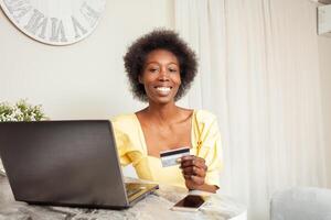 schön afrikanisch amerikanisch schwarz Frau Verwendet ein Bank Karte zu Einkaufen online. Lieferung von Waren zu Haus. Laptop auf Tisch. lächelnd, glücklich foto