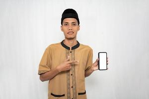 asiatisch Muslim Mann tragen braun Muslim Kleider lächelnd glücklich während zeigen beim das Smartphone Bildschirm. isoliert Weiß Hintergrund. foto