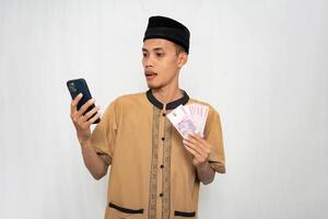 asiatisch Muslim Mann tragen Muslim Kleider ist schockiert suchen beim das Smartphone Bildschirm während Tragen Geld im seine andere Hand. isoliert Weiß Hintergrund. foto