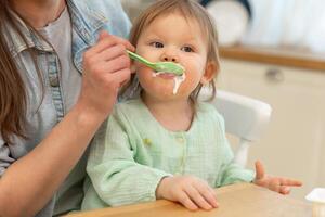 glücklich Familie beim heim. Mutter Fütterung ihr Baby Mädchen von Löffel im Küche. wenig Kleinkind Kind mit unordentlich komisch Gesicht isst gesund Essen beim heim. jung Frau Mama geben Essen zu Kind Tochter foto