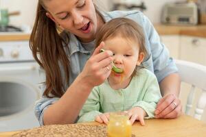 glücklich Familie beim heim. Mutter Fütterung ihr Baby Mädchen von Löffel im Küche. wenig Kleinkind Kind mit unordentlich komisch Gesicht isst gesund Essen beim heim. jung Frau Mama geben Essen zu Kind Tochter foto