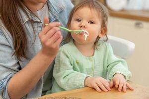 glücklich Familie beim heim. Mutter Fütterung ihr Baby Mädchen von Löffel im Küche. wenig Kleinkind Kind mit unordentlich komisch Gesicht isst gesund Essen beim heim. jung Frau Mama geben Essen zu Kind Tochter foto