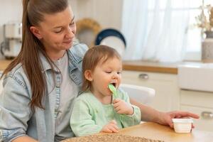 glücklich Familie beim heim. Mutter Fütterung ihr Baby Mädchen von Löffel im Küche. wenig Kleinkind Kind mit unordentlich komisch Gesicht isst gesund Essen beim heim. jung Frau Mama geben Essen zu Kind Tochter foto