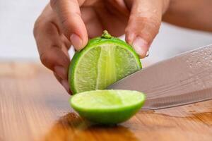 weiblich Hand halten Messer ist Schneiden Limette auf ein hölzern Tafel im Küche. Schneiden frisch Zitrone im Hälfte schließen hoch. Höhe Vitamin c natürlich foto