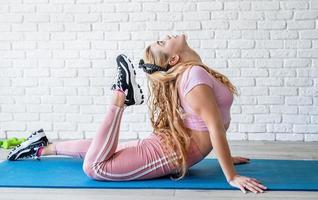 athletische Frau, die sich zu Hause auf einer Fitnessmatte am weißen Backsteinmauerhintergrund ausdehnt foto