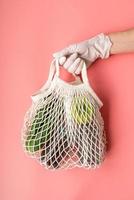 Hand in einem Handschuh, der eine weiße, umweltfreundliche Netztasche mit Lebensmittelvorräten auf rosafarbenem Hintergrund hält
