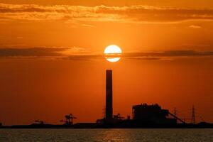 Sonnenuntergang hinter ein industriell Fabrik und im Vorderseite ist das Meer, Orange Farbe, Silhouette foto