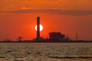 Sonnenuntergang hinter ein industriell Fabrik und im Vorderseite ist das Meer, Orange Farbe, Silhouette foto