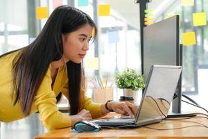 Junge asiatische Programmiererin in einem gelben Hemd steht nach unten, um ihren Laptop und PC im Büro zu benutzen.