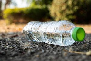 Nahaufnahme klares Plastikflaschenwassergetränk mit grüner Kappe auf der Straße im Park bei unscharfem Hintergrund, Müll, der außerhalb der Tonne zurückgelassen wird foto