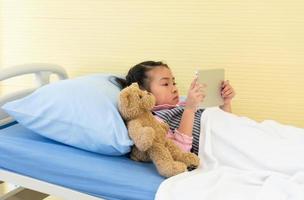 junges asiatisches mädchen liegt im krankenhausbett und sieht sich cartoon in tablette an. Gesundheits- und Medizinkonzept foto