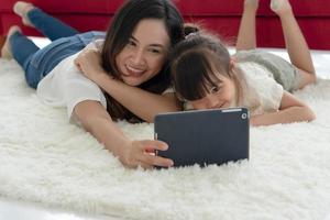 asiatisches Kind, das ihre Mutter küsst, während die Mutter mit Tablet ein Foto von ihnen im Wohnzimmer macht. Familie zusammen Konzept