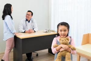 junges asiatisches mädchen, das teddybär hält und vor mutter und arzt in der krankenhausentwicklungsklinik steht. Gesundheits- und Medizinkonzept foto