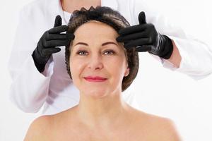 Plastischer Chirurg, der das Gesicht der Frau mittleren Alters vor der Schönheitsoperation überprüft, Kollagen, Falten foto
