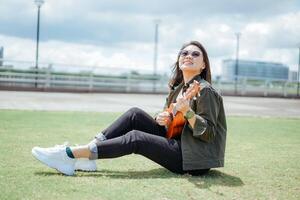 spielen Ukulele von jung schön asiatisch Frau tragen Jacke und schwarz Jeans posieren draußen foto