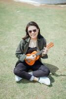 spielen Ukulele von jung schön asiatisch Frau tragen Jacke und schwarz Jeans posieren draußen foto