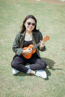 spielen Ukulele von jung schön asiatisch Frau tragen Jacke und schwarz Jeans posieren draußen foto