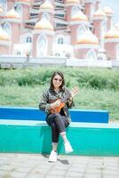 spielen Ukulele von jung schön asiatisch Frau tragen Jacke und schwarz Jeans posieren draußen foto