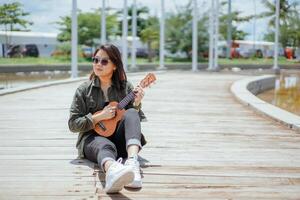 spielen Ukulele von jung schön asiatisch Frau tragen Jacke und schwarz Jeans posieren draußen foto