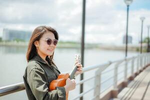 spielen Ukulele von jung schön asiatisch Frau tragen Jacke und schwarz Jeans posieren draußen foto