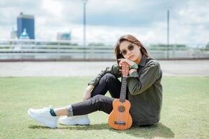 spielen Ukulele von jung schön asiatisch Frau tragen Jacke und schwarz Jeans posieren draußen foto