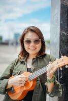 spielen Ukulele von jung schön asiatisch Frau tragen Jacke und schwarz Jeans posieren draußen foto