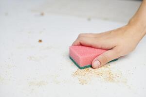 Schwamm in Frauenhand entfernt Schmutz, Semmelbrösel und Essensreste. Küchentisch reinigen. Hausarbeiten foto