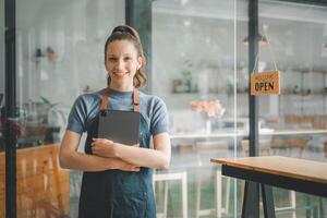 schön jung Barista Frau im Schürze halten Tablette und Stehen im Vorderseite von das Tür von Cafe mit öffnen Zeichen Tafel. Geschäft Inhaber Start-up, sme Unternehmer Verkäufer Geschäft Konzept. foto