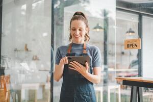 schön jung Barista Frau im Schürze halten Tablet und Stehen im Vorderseite von das Tür von Cafe mit öffnen Zeichen Tafel. Geschäft Inhaber Anfang Konzept. foto