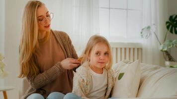 kaukasisch Mutter Kamm Haar von süß preteen Kind Mädchen Tochter Mama Babysitter Hilfe wenig Kind Kind Baby mit Bürsten Kämmen Frisur Frisur bereiten Schönheit Routine Sitzung beim Zuhause auf Couch Sofa foto