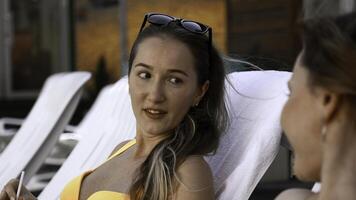 glücklich jung Frauen Lügen auf Sonnenliegen durch das Schwimmen Schwimmbad und Klatschen. Clip. entspannend in der Nähe von Resort Schwimmen Schwimmbad mit ein Cocktail. foto
