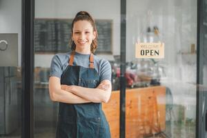 Porträt von glücklich Kellnerin Stehen beim Restaurant Eingang mit öffnen Zeichen, Porträt von jung Geschäft Frau besuchen Neu Kunden im ihr Kaffee Geschäft. foto