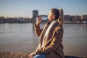 Frau genießt Trinken Wasser während Sitzung durch das Fluss auf ein sonnig Winter Tag. getönt Bild. foto