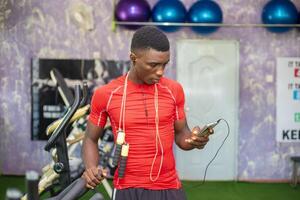 gut aussehend afrikanisch Kerl im das Fitnessstudio Betriebs seine Handy und Hör mal zu zu Musik- foto